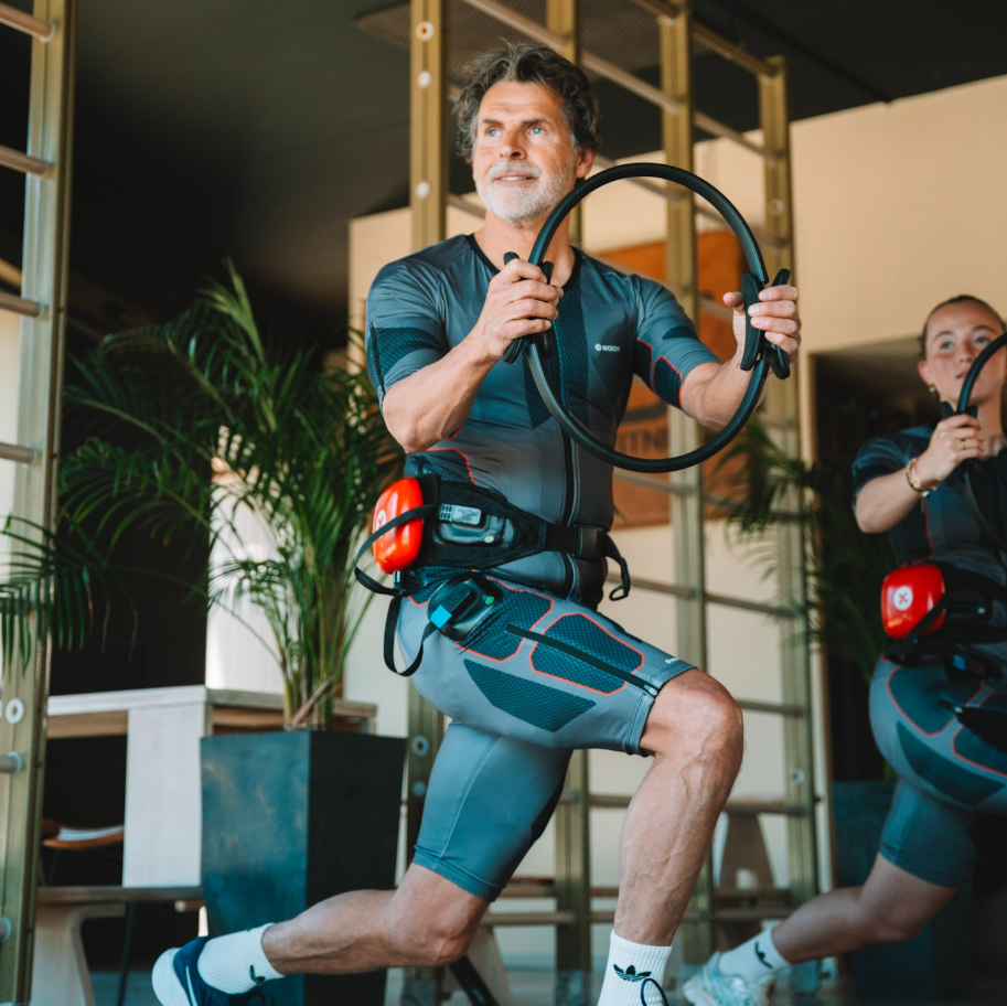 Moderne EMS-studio met trainer en ondernemer bij draadloze trainingsapparatuur, pakken en tablet in een premium ruimte met warme lichte accenten.