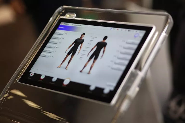 Trainer stelt een draadloos EMS apparaat in naast een sporter in EMS-pak, in een moderne premium studio met zacht daglicht en strakke apparatuur.